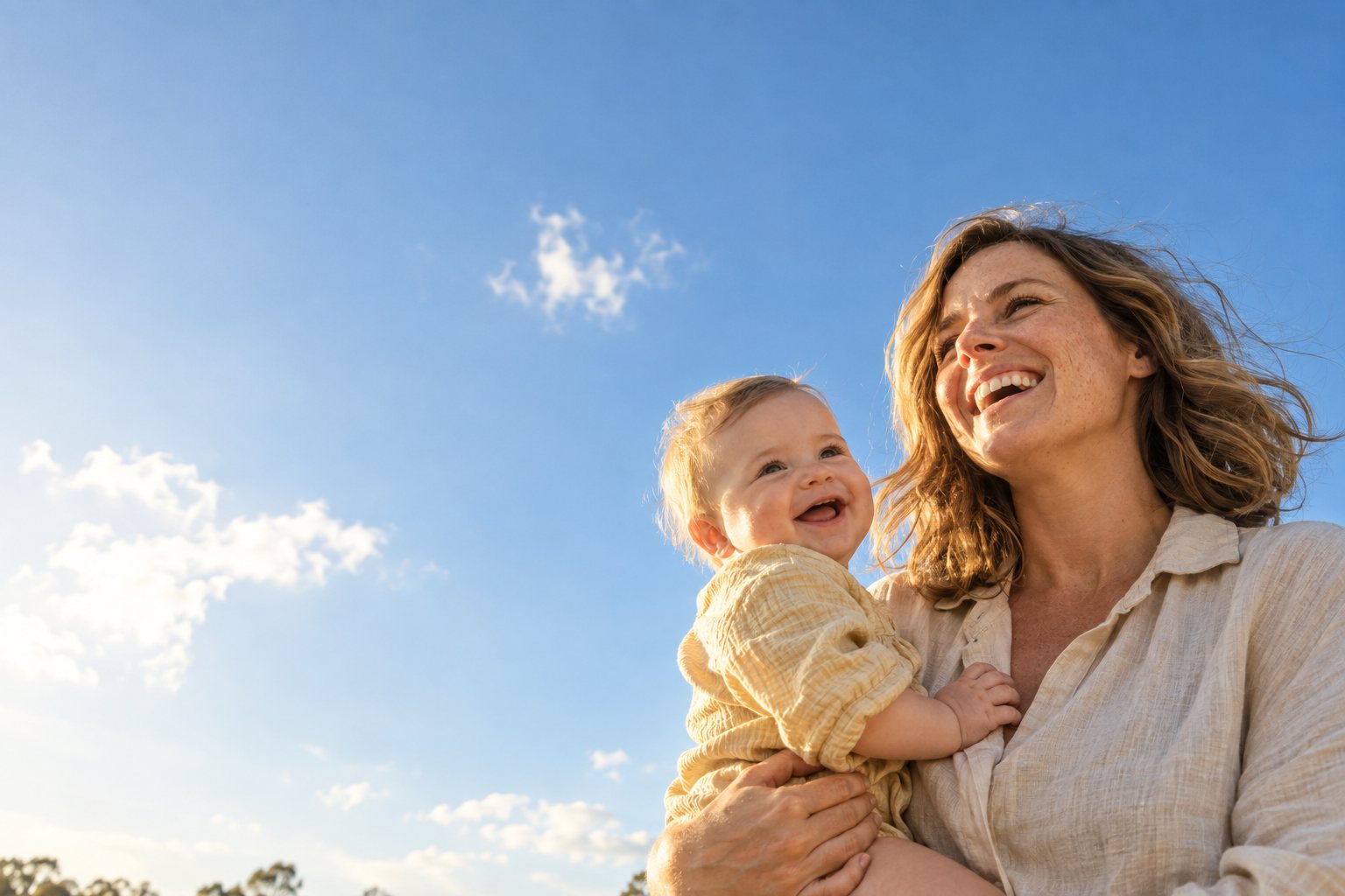 Sarah laughs in the sunshine, holding baby Charlie up against a clear blue sky